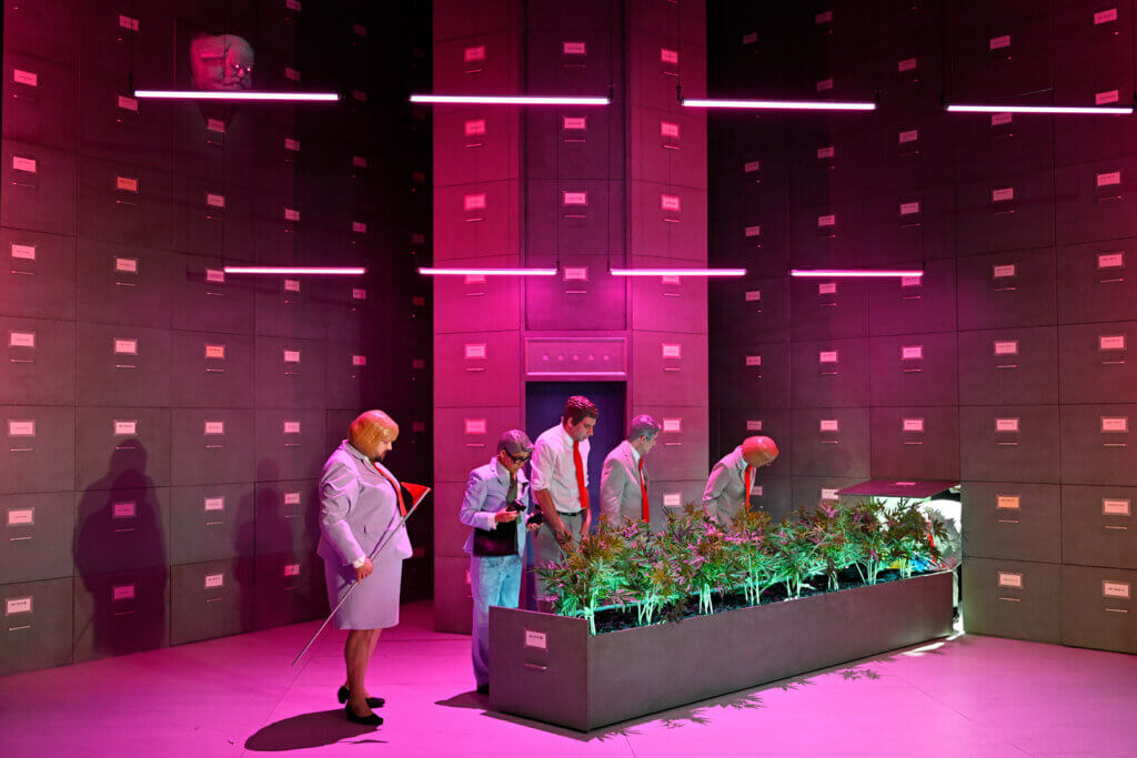 Aufführungsfoto von „Wie Grischa mit einer verwegenen Idee beinahe den Weltfrieden auslöste“ am Deutschen Theater Berlin. In einem rot erleuchteten Büroraum stehen fünf Angestellte vor einer Schublade, aus der Pflanzen wachsen.