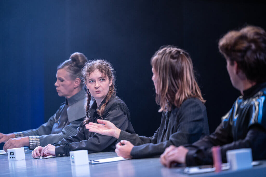Aufführungsfoto von „√Mytopia: Auferstanden aus Ruinen“ am Theater Altenburg Gera. Drei Frauen und ein Mann sitzen an einem Tisch nebeneinander.