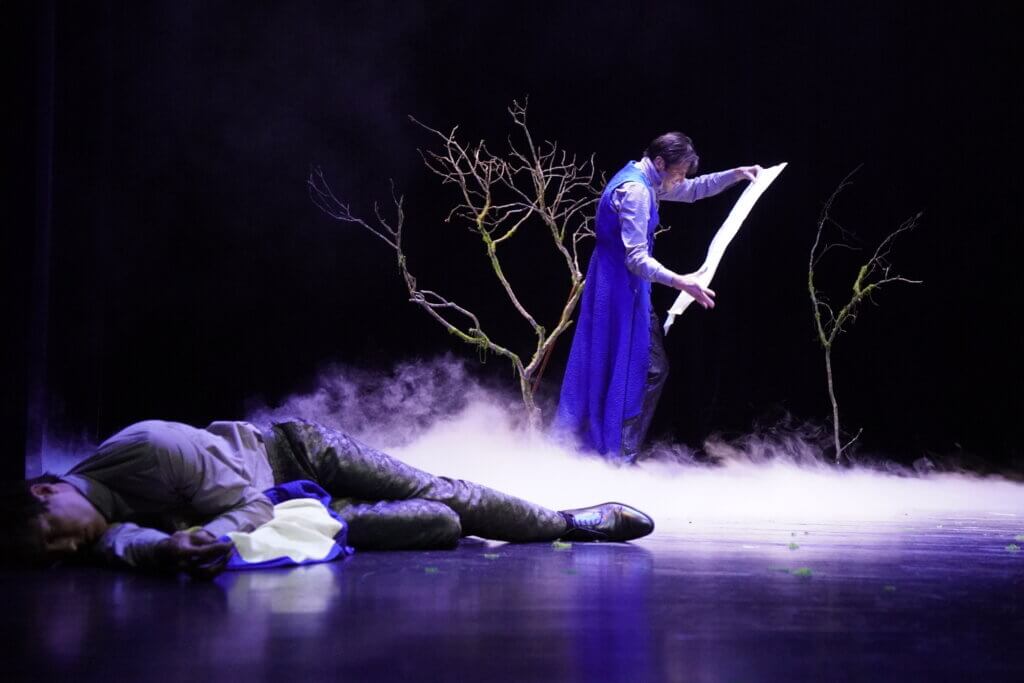Aufführungsfoto von „Faust. Eine Tragödie“ am Landestheater Schwaben. Ein Mann liegt links am Boden, ein anderer Mann steht im Hintergrund vor einem Baum und schaut sich ein langes weißes Papier an.