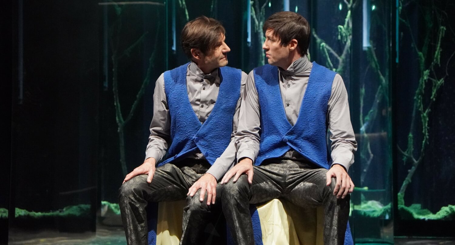 Aufführungsfoto von „Faust. Eine Tragödie“ am Landestheater Schwaben. Zwei sehr ähnlich aussehende Männer, die gleich gekleidet sind, sitzen nebeneinander und schauen sich an.