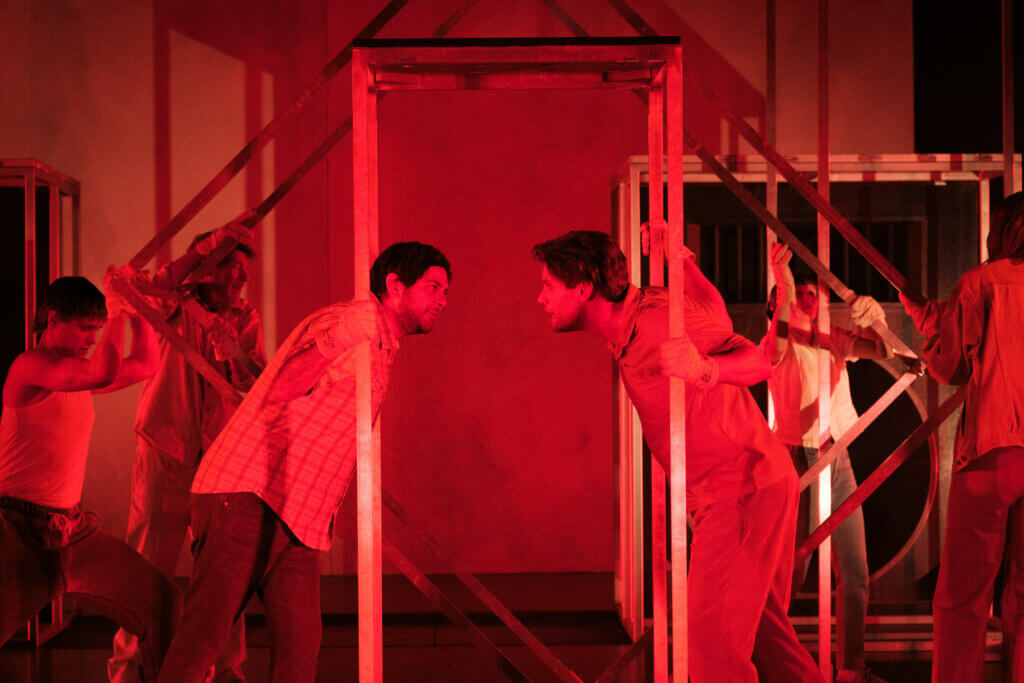 Aufführungsfoto von „Jakob der Lügner“ am Stadttheater Gießen. Die Bühne ist in warm-rotes Licht getaucht. Zwischen einem offenen Stahl-Rechteck stehen sich zwei Männer gegenüber und schauen sich an.