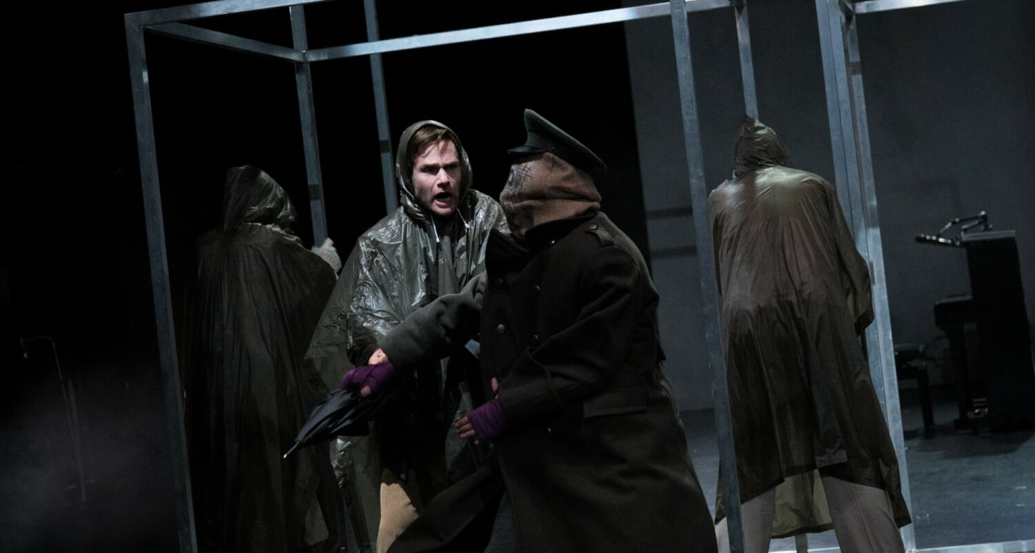 Aufführungsfoto von „Jakob der Lügner“ am Stadttheater Gießen. Drei Personen mit dunklen Regencapes, einer im Vordergrund schreit eine Gestalt an mit schwarzen Mantel, Offiziersmütze und unkenntlichem Gesicht.