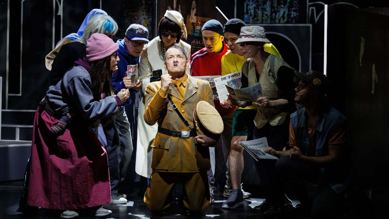 Aufführungsfoto von „Er ist wieder da“ von Jack Woodhead am Theater für Niedersachsen. Ein Mann, der wie Adolf Hitler aussieht kniet in der Mitte, um ihn herum schauen ihn andere Personen a und halten Zeitungen in den Händen.
