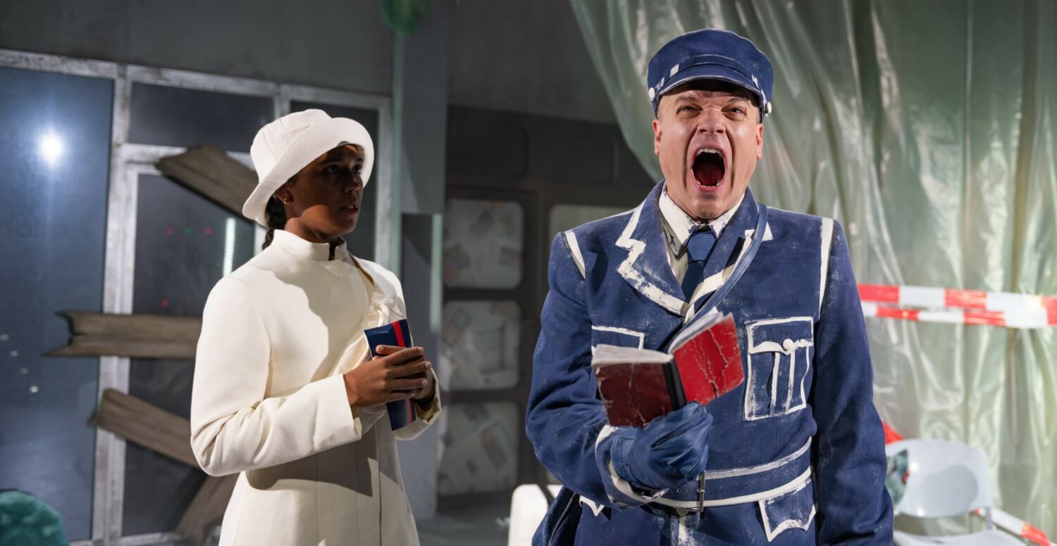 Aufführungsfoto von „Das Deutschlandherz“. Ein Mann in blau-weißer Uniform und einem roten Notizbuch in der Hand reißt den Mund auf. Eine Frau in einem weißen Kostüm mit weißem Hut steht daneben.