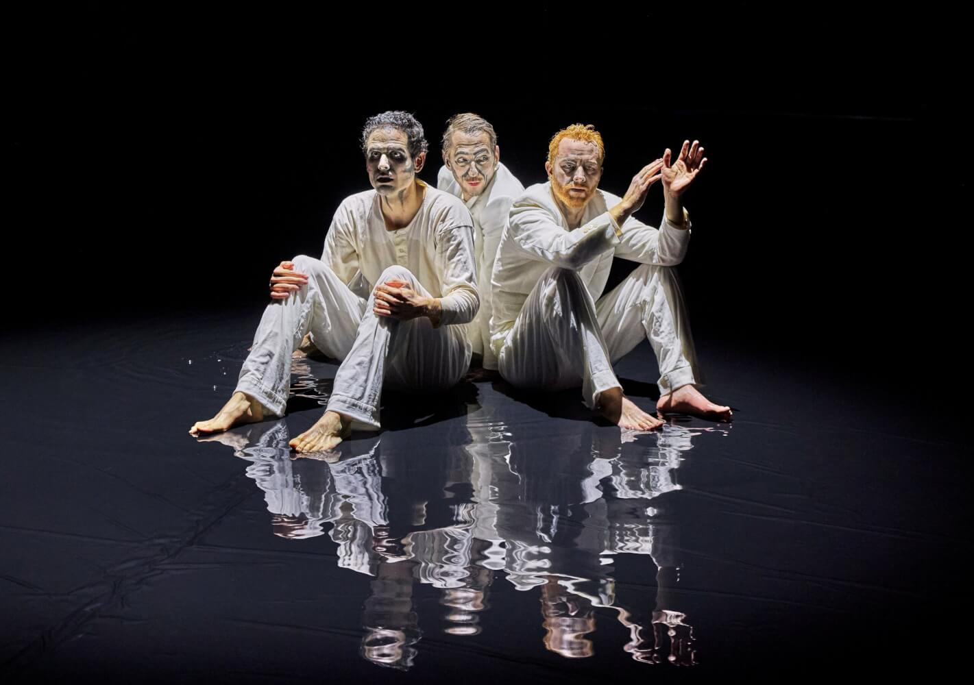 Leon Schamlott, Hannes Berg und Loris Kubeng sitzen in der Mitte der Bühne auf dem Boden, die Beine angewinkelt. Der Boden spiegelt ihre Formation.