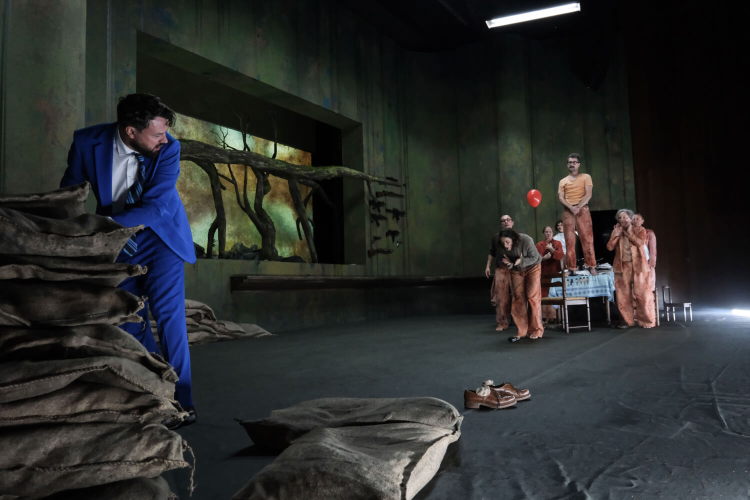 Aufführungsfoto von „Catarina oder von der Schönheit, Faschisten zu töten“ von Tiago Rodrigues am Theater Bremen. Ein Mann im blauen Anzug steht links hinter einem Haufen Sandsäcke, rechts um einen Tischgruppiert stehen andere Menschen, die im zuschauen.
