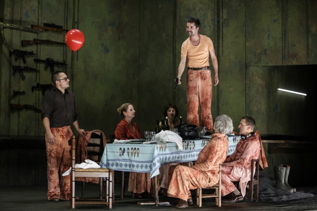 Aufführungsfoto von „Catarina oder von der Schönheit, Faschisten zu töten“ am Theater Bremen. Ein Mann steht auf einem Tisch und hält eine Pistole nach unten. Um den Tisch sitzen vier Menschen, eine Person steht.