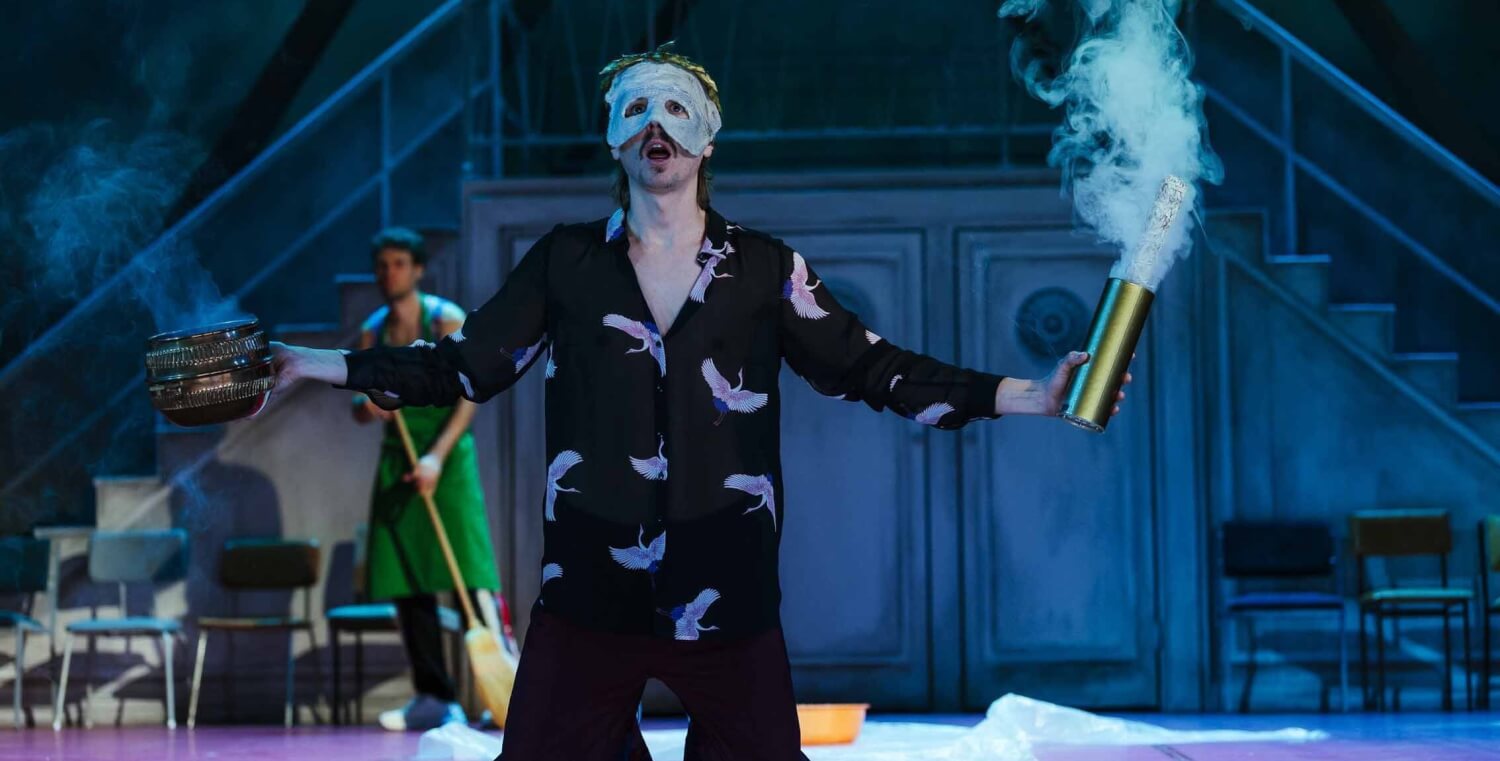 „Der Frieden“ von Peter Hacks am Staatstheater Cottbus. Ein Mann kniet auf dem Boden. Er hat eine weiße Maske auf und hält in den Händen zwei Gefäße, aus denen Rauch aufsteigt.