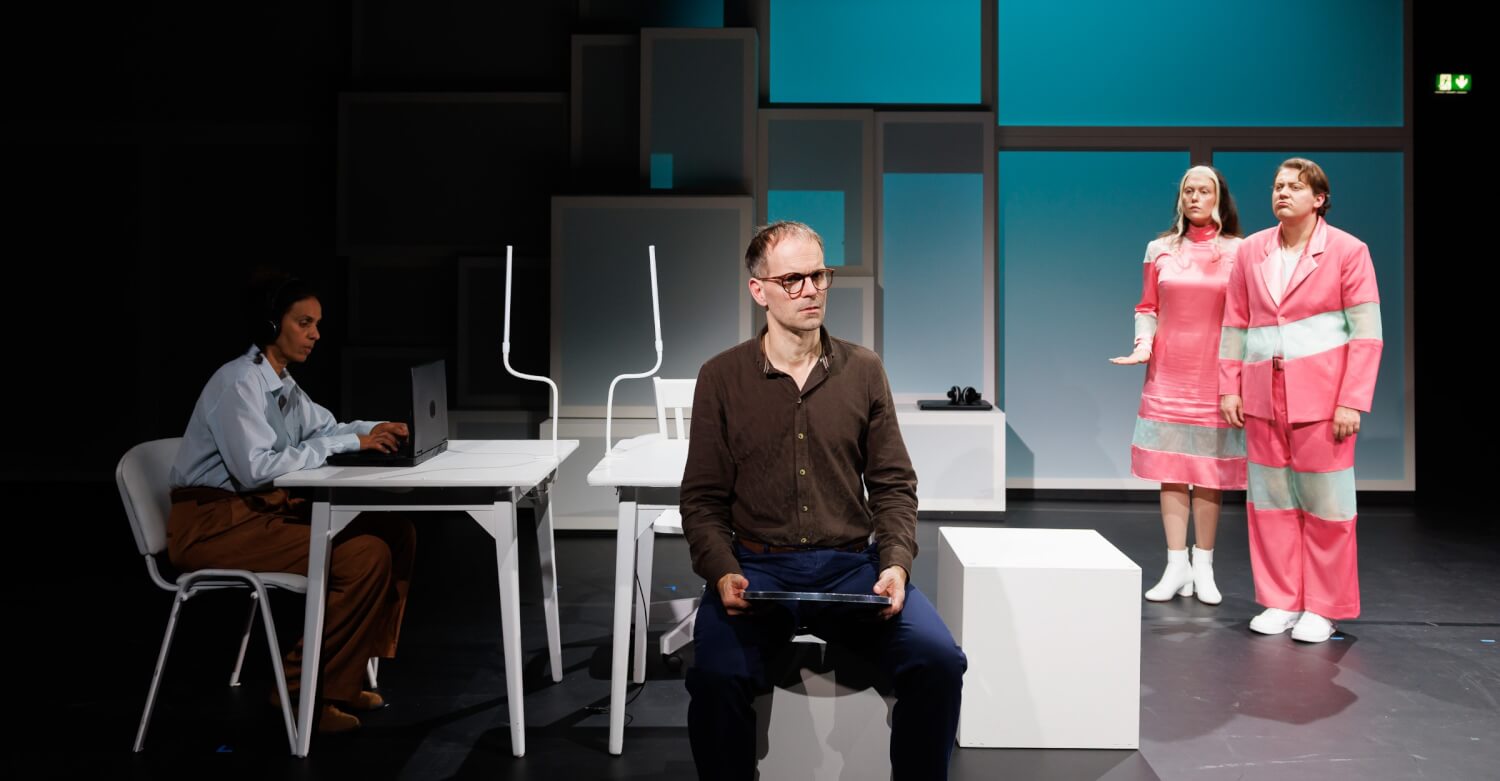 Aufführungsfoto von „Der öffentliche Raum“ am Hessischen Landestheater Marburg. Ein Mann sitzt im Vordergrund mit einem Tablet in der Hand. Links sitzt eine Frau am Laptop. Rechts stehen zwei Personen.