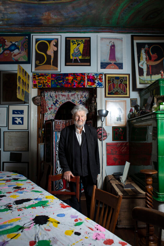 Achim Freyer im Kunsthaus der Achim Freyer Stiftung. Er steht hinter einem Stuhl und hält sich an der Lehne fest, hinter ihm an der Wand viele verschiedene Bilder und vor ihm ist der Tisch mit einer bunt beklecksten Tischdecke überzogen.