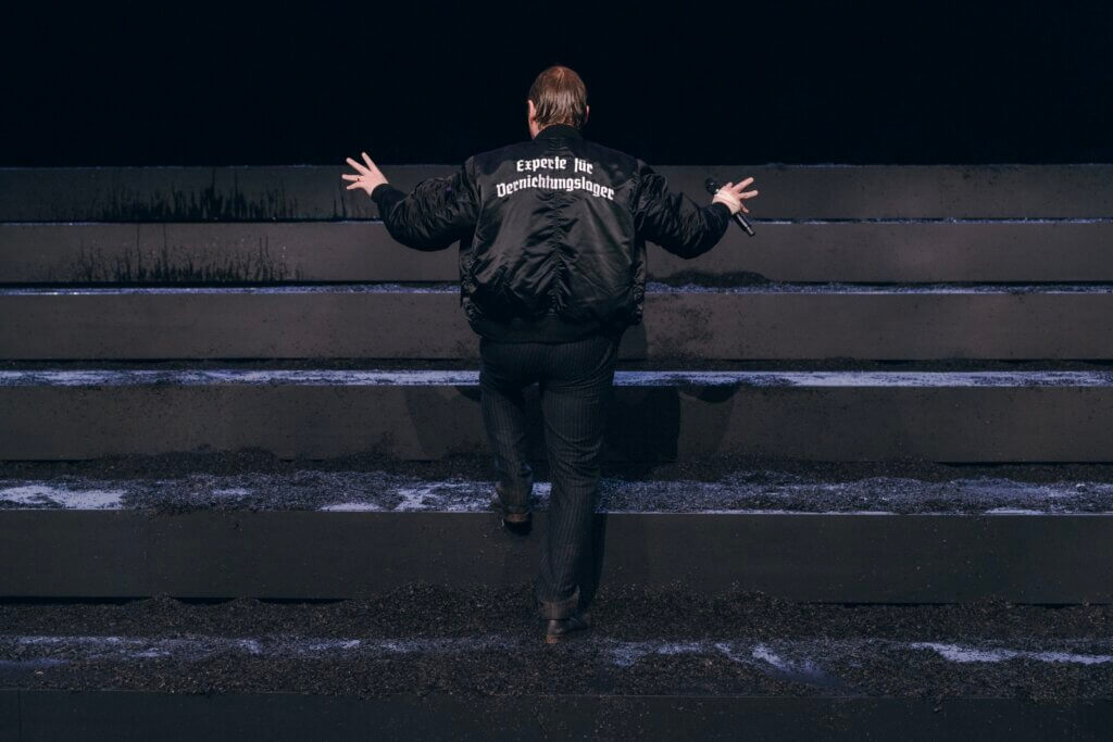 eine Person läuft auf einer Theaterbühne eine Treppe hoch mit ausgestreckten Armen und einer schwarzen Jacke mit der Aufschrift „Experte für Vernichtungslager“