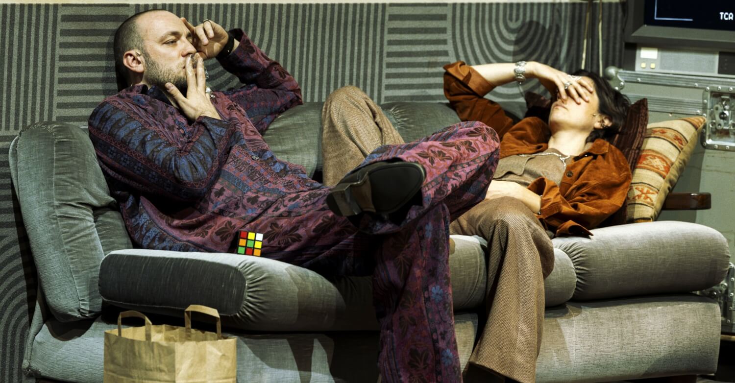 Aufführungsfoto von „Stereophonic“ von David Adjmi an der Schaubühne Berlin. Ein Mann und eine Frau liegen auf einer Couch. Er raucht eine Zigarette, die bedeckt ihre Augen mit ihrer rechten Hand.