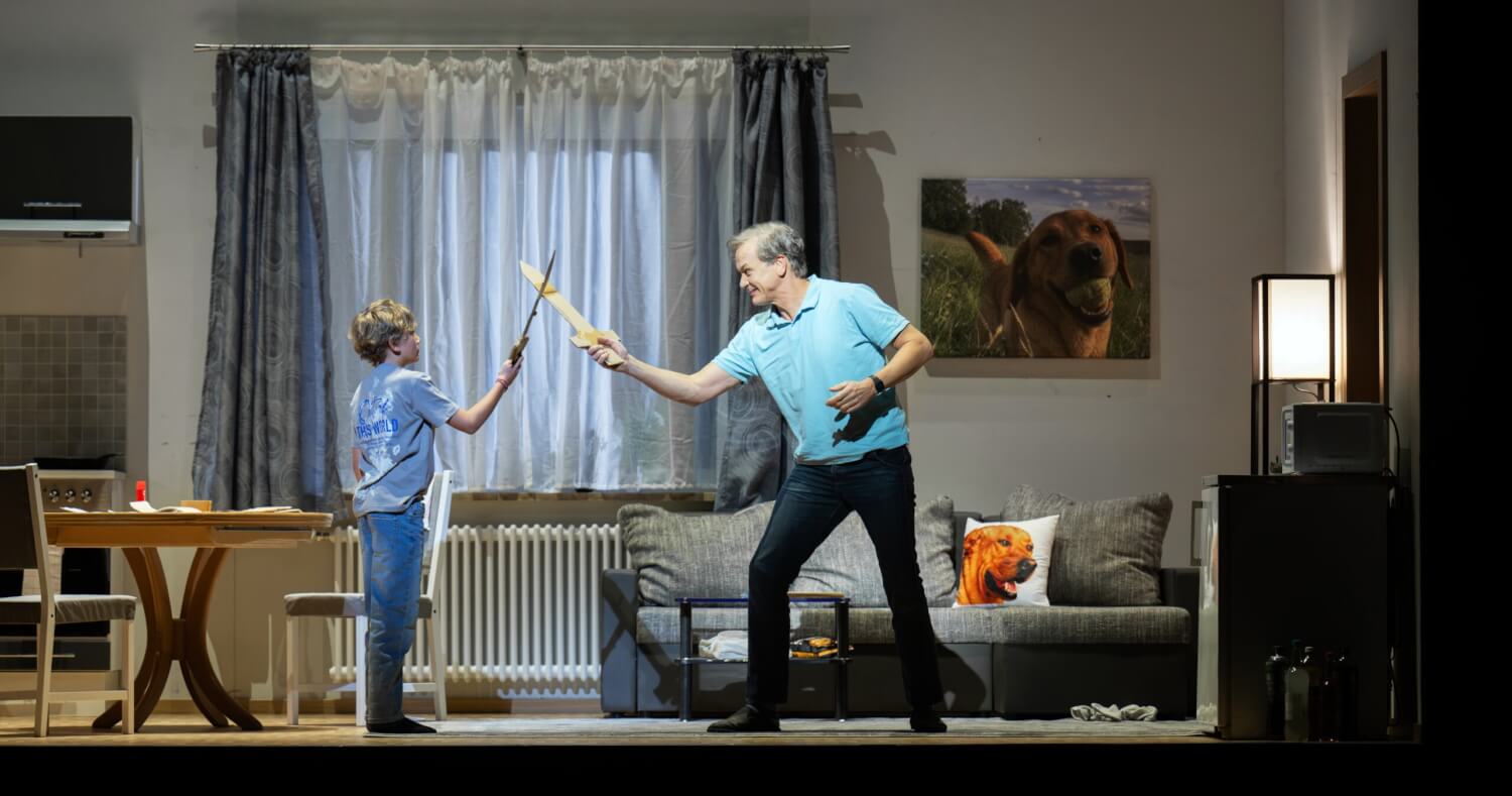 „Siegfried“ von Richard Wagner an der Bayerischen Staatsoper. Ein Junge und ein Mann stehen sich in einem Wohnzimmer mit Holzschwertern gegenüber und kämpfen.