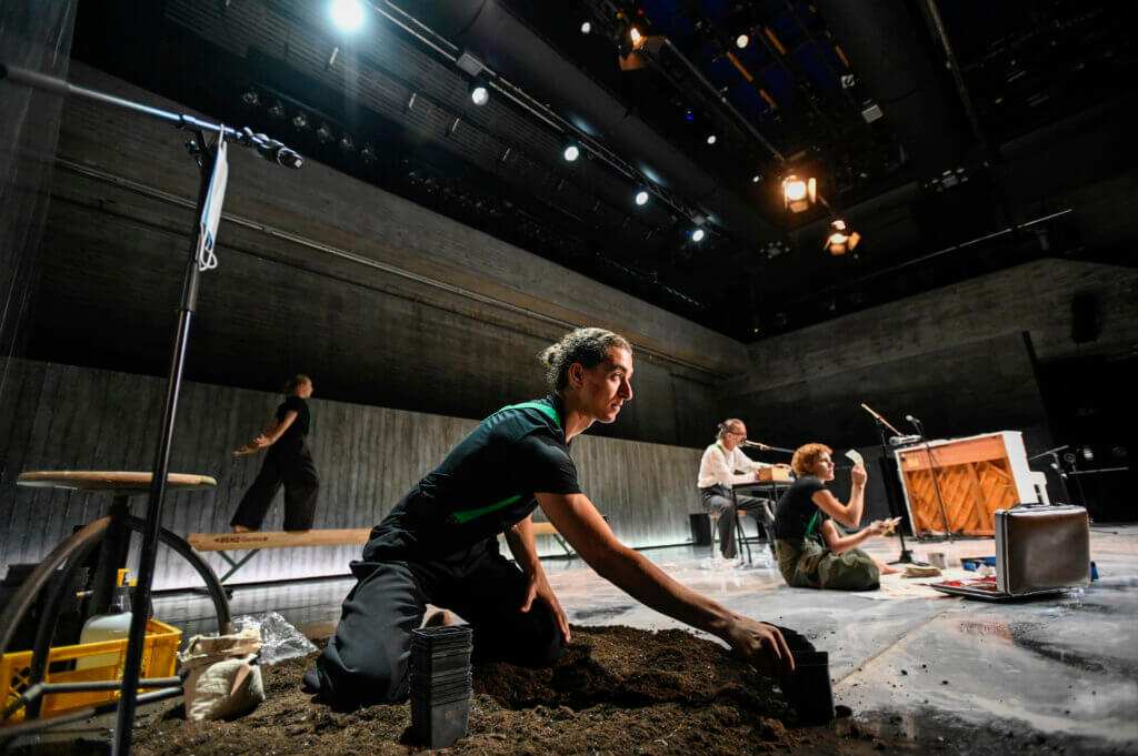 Aufführungsfoto von „Das Prinzip Hoffnung“ am Badischen Staatstheater Karlsruhe. Ein Mann kniet vorne in Erde. Im Hintergrund läuft eine Frau auf einem Schwebebalken, ein Mann sitzt mit einem Mikrofon an einem Tisch, eine andere Frau schaut sich ein kleines Foto hockend an.