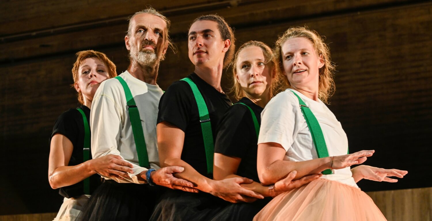 Aufführungsfoto von „Das Prinzip Hoffnung“ am Badischen Staatstheater Karlsruhe. Fünf Personen steht dicht hintereinander aufgereiht, alle tragen grüne Hosenträger. Ihre Arme berühren sie mit ihren Händen.