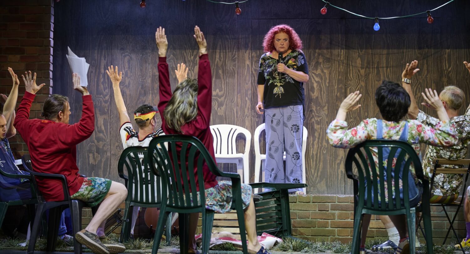 „Zur fröhlichen Zwietracht!“ am Hans Otto Theater Potsdam. Mehrere Personen sitzen auf Gartenstühlen und heben die Hände in die Höhe. Eine Frau steht ihnen gegenüber mit einem Mikro in der Hand und schaut sie unsicher an.