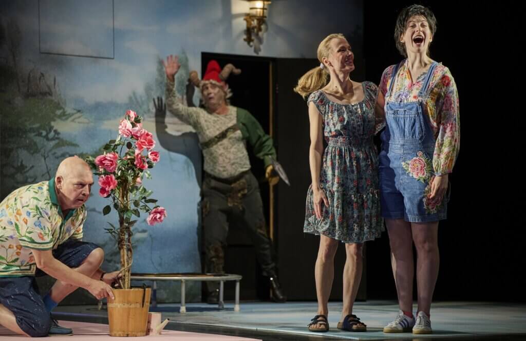 „Zur fröhlichen Zwietracht!“ am Hans Otto Theater Potsdam. Zwei Frauen steht nebeneinander, links daneben versteckt sich ein älterer Mann kniend hinter einem Rosenstock. Im Hintergrund winkt ein Mann mit einem Steinbockhut auf dem Kopf.